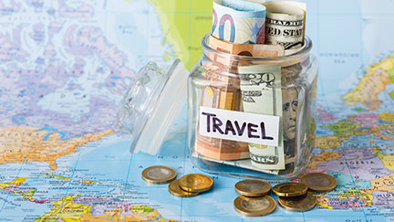 Un bote de cristal, con una pegatina en la que podemos leer "travel", lleno de billetes de distinos paises, junto a unas monedas, sobre un mapa del mundo